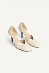 Zapato de tacón de piel color beige, con detalle cruzado en la parte delantera y tacón alto fino combinado con acabado en punta redonda.