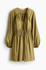 Vestido corto oversized de mezcla de viscosa y color verde militar, con escote en V profundo, cordón ajustable en el cuello y mangas globo largas con puños elásticos. Presenta fruncidos en el canesú y sección de punto smock en la cintura con falda de leve vuelo.