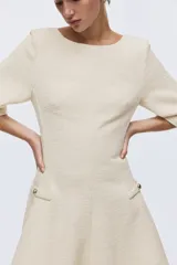 Vestido corto de tejido bouclé color beige, con escote redondo, espalda en V y mangas francesas. Cuenta con bolsillos decorativos con botones dorados en la cintura y falda con vuelo.