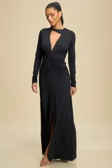 Vestido largo de manga larga en color negro, confeccionado en tela elastizada. Presenta un diseño con escote profundo y detalle de abertura superior, con drapeado central en la cintura que estiliza la silueta y una abertura frontal en la falda.