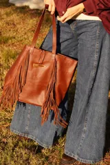 Bolso tipo tote de cuero color marrón, con diseño de flecos laterales y cierre frontal mediante un botón de guampa. Cuenta con doble asa de hombro.