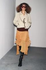 Trench corta color beige con cuello solapa, manga larga con trabilla y botón en los puños, bolsillos de vivo en el delantero y cierre frontal con botones oscuros.