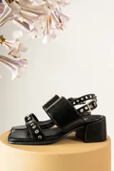 Sandalias de gamuza color marrón con taco cuadrado de madera. Presentan dos tiras anchas, una superior lisa y una inferior decorada con ojales metálicos plateados. Cuentan con cierre de pulsera con hebilla ajustable en el tobillo.