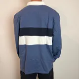 Buzo tipo polo de rugby con cuello blanco, manga larga y puños acanalados. Presenta un diseño de bloques de color en azul marino y verde, con un pequeño logo bordado en el pecho.