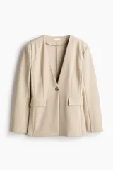 Blazer de punto grueso en color beige, con diseño sin cuello, escote en V, hombreras y cintura entallada. Cuenta con cierre de un botón frontal y bolsillos delanteros con solapa.