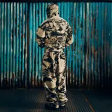 Campera con capucha de corte holgado, confeccionada en tela con estampado camuflado en tonos grises. Presenta un diseño gráfico bordado en la espalda que incluye un águila central, texto y detalles numéricos.