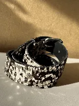 Cinturón ancho de cuero con estampado de vaca (blanco y negro) y múltiples tachas metálicas redondas. Incluye una hebilla metálica y una tachuela piramidal como detalle central.