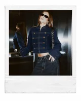 Chaqueta de denim azul oscuro con estilo militar, cuello alto, cierre frontal con cremallera y detalles de trabas decorativas con botones dorados.