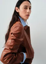 Chaqueta de cuero ovino color marrón, con diseño corto, cuello camisero, cierre frontal con botones y puños elásticos.