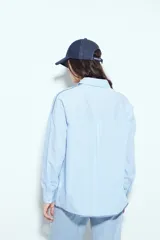 Camisa de silueta amplia y caída fluida, color celeste con rayas verticales blancas. Presenta cuello camisero clásico, cierre frontal con botones y un bolsillo aplicado en el pecho. Las costuras están resaltadas en azul oscuro.