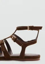 Sandalias planas de gamuza marrón con diseño de múltiples tiras cruzadas y hebillas metálicas doradas. Cuenta con cierre de hebilla en el tobillo.