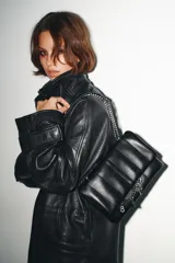 Bolso bandolera de diseño acolchado en color negro, con solapa frontal decorada con una cadena metálica y charms colgantes. Cuenta con cierre de botón imantado, triple compartimento interior y correa ajustable que permite llevarlo al hombro o cruzado.