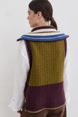 Chaleco de lana con cierre frontal completo y cuello alto. Presenta un diseño de bloques de color en tonos marrón chocolate y celeste, combinando secciones de tejido trenzado con secciones de tejido acanalado. Posee un corte asimétrico, siendo más largo en la parte posterior, y aberturas laterales en el ruedo.