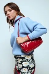 Cartera de hombro de diseño rectangular, confeccionada en material sintético con acabado brillante. Presenta costuras decorativas verticales y cierre metálico en el compartimento principal. Incluye una correa fija con detalles de ojalillos metálicos.