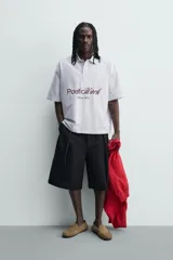 Camisa polo boxy fit de manga corta, confeccionada en tejido de algodón con diseño de rayas verticales celestes y blancas. Cuenta con cuello solapa, cierre frontal con botones y estampado frontal de texto en tono marrón con la leyenda "Poetic Work Since 1975".