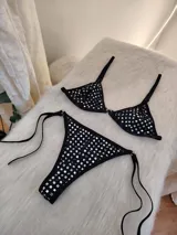 Conjunto de bikini negro con apliques de tachas cuadradas plateadas. El corpiño es de estilo triangular con una argolla central metálica y breteles regulables. La bombacha es de corte colaless con tiras laterales ajustables y argollas metálicas en los extremos.