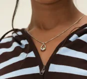 Collar de cadena fina plateada con un dije colgante de metal plateado con forma ovalada y texturizada, similar a un candado abierto o una piedra pulida.