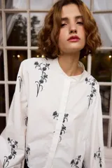 Camisa de algodón color blanco con cuello mao y cierre frontal de botones. Presenta un diseño de bordados florales en color negro distribuidos en el frente y las mangas. Posee mangas largas abullonadas con puños abotonados y un corte recto ligeramente cropped.