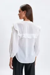 Camisa blanca de corte casual con cuello redondo, cierre frontal de botones y detalle de volados en los hombros. Presenta mangas tres cuartos con puños elásticos.