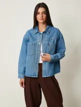 Camisa de denim celeste oversize con bolsillos frontales, cierre de botones metálicos y detalles de tachas en el cuello y la parte delantera.