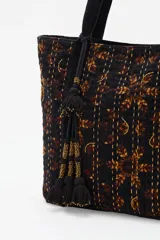 Bolso tote de algodón negro con estampado en tonos marrones y ocres, con costuras visibles en color claro. Presenta tiras de hombro negras y un adorno colgante con borlas negras y detalles dorados.