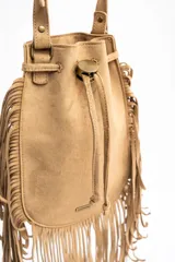 Cartera tipo bucket bag color camel, confeccionada en material sintético con acabado gamuzado. Presenta un diseño decorado con flecos largos en todo el contorno, cierre mediante cordón ajustable y correa larga regulable.