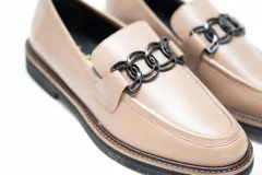 Mocasín de cuero vacuno color visón, con diseño clásico de punta redondeada y costuras visibles. Presenta un adorno metálico negro en el empeine y suela plana de goma negra.
