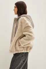 Campera de estilo híbrido con cuerpo de corderito sintético color beige y panel frontal central con cierre y capucha de tela técnica metalizada en tono grisáceo. Presenta cordones ajustables en la capucha con diseño estampado y puños con detalle de rayas.