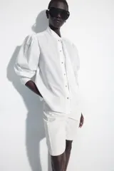 Camisa de algodón color blanco, con cuello subido y detalle de pañuelo incorporado, manga larga con puños anchos y cierre frontal con botones joya dorados.