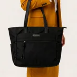 Cartera tipo tote bag de color negro con diseño estructurado, asas largas de hombro en cuero sintético, bolsillo frontal con cierre y placa metálica con el logo de la marca.