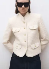 Chaqueta de tweed color crema con cuello mao, cierre frontal de botones dorados efecto joya y cuatro bolsillos delanteros con solapa. Presenta diseño recto, manga larga y forro interior.