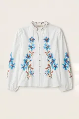 Camisa blanca de manga larga abullonada, con delicados bordados florales en tonos azules y verdes a lo largo de la botonadura frontal y en las mangas. Presenta cuello mao con reborde en contraste y cierre frontal con botones.