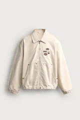 Cazadora de algodón color beige con efecto lavado, corte relaxed fit, cuello solapa y cierre frontal con cremallera. Cuenta con bolsillos frontales tipo canguro, puños y bajo elásticos, y aplicaciones de parches bordados a contraste en el delantero.