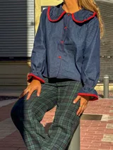 Camisa de jean azul oscuro con cuello estilo marinero y puños con volados, ambos con ribetes en color rojo. Presenta cierre frontal con botones rojos.
