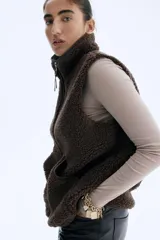 Chaleco de sherpa color marrón con cuello elevado, cierre frontal y bolsillos tipo parche delanteros. Presenta ribetes de cinta y forro interior.