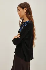 Sweater negro de punto con cuello redondo y mangas largas. Presenta una aplicación decorativa de lentejuelas y pedrería en tonos azules y oscuros sobre el hombro izquierdo.