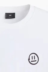 Remera blanca de punto de algodón de peso medio, con cuello redondo ribeteado y hombros caídos. Presenta un pequeño estampado minimalista de un emoji con la boca recta en el pecho.