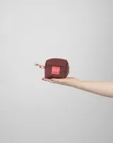 Monedero pequeño de tela color bordó con cierre superior y gancho metálico lateral. Presenta una etiqueta rectangular con el logo de la marca en el frente.
