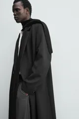 Abrigo largo de corte relajado en color negro, confeccionado en tejido con viscosa. Presenta un diseño de botonadura cruzada, cuello con solapas de pico, manga larga con puños abotonados y bolsillos laterales de vivo. Incluye una abertura en el centro de la espalda y un bolsillo interior.