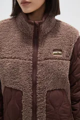 Campera de diseño híbrido que combina paneles de corderito sintético en color crema con mangas y bolsillos de tela técnica acolchada en color verde militar. Presenta cierre frontal completo, cuello alto y un pequeño parche decorativo en el pecho.