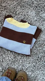Buzo tejido de punto con diseño de rayas horizontales en color marrón chocolate y celeste, con cuello redondo en color amarillo y detalle de pequeño bordado en el pecho.