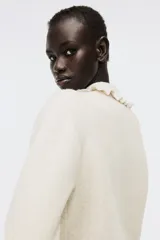 Saco corto de punto suave color beige, con cuello camisero con ribete de volados y botones de metal con diseño de nudo. Tiene hombros ligeramente caídos y remate acanalado en puños y bajo.