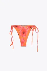 Braguita de bikini estampada con flores rojas y anaranjadas sobre fondo naranja. Tiene tiro medio y cintura ajustable con lazadas.