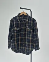 Camisa de franela a cuadros en tonos negro y marrón, con cuello clásico, cierre frontal de botones, mangas largas con puños y dos bolsillos frontales con solapa.