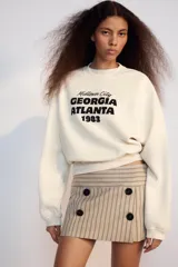 Falda pantalón de tiro bajo color beige con diseño de rayas diplomáticas verticales en tonos oscuros, con detalles de cuatro botones negros decorativos en la parte frontal y cierre lateral con cremallera oculta.