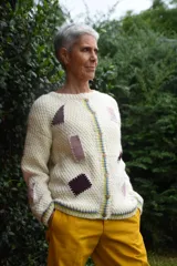 Cardigan de lana merino tejido a mano con técnica de dos agujas, de color marfil con parches geométricos de tela en tonos celestes y lila aplicados con costuras visibles. Presenta cierre frontal con botones de madera y detalles de ribetes multicolores en los bordes.