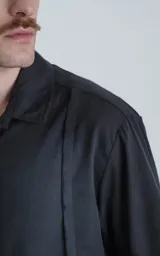 Camisa de manga corta en color negro con proceso de lavado, cuello clásico y cierre frontal con botones. Presenta un diseño minimalista con una costura vertical decorativa en el frente.