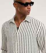 Camisa de manga larga con cuello inglés y cierre frontal de botones. Presenta un diseño de rayas verticales en tonos neutros, confeccionada en tejido de poliéster con caída estructurada y corte comfort.
