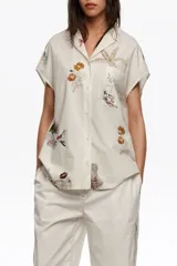 Camisa de manga corta color blanco con cuello solapa y estampado de temática marina con peces y estrellas de mar, con cierre frontal de botones y un bolsillo en el pecho.