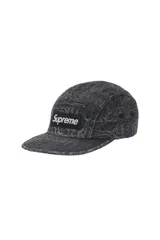 Gorra tipo camp cap de Supreme, confeccionada en denim con efecto lavado y estampado de estilo gótico en toda la superficie. Presenta el logo de la marca en un parche rectangular frontal y ojales laterales para ventilación.
